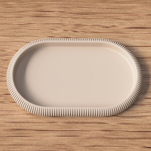 Puede incluir: Una bandeja ovalada beige con borde acanalado, sobre un fondo de madera. El dise&ntilde;o de la bandeja es sencillo y elegante, adecuado para servir o exhibir. La bandeja es de color neutro.