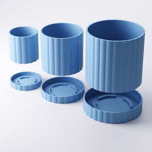 May include: Three blue cylindrical planters of varying sizes, each with a matching saucer. The planters have a ribbed design and are arranged against a white background. The largest planter is approximately 7 inches tall and 6 inches in diameter.