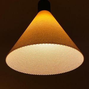 May include: A cone-shaped pendant light fitting with a textured, off-white exterior surface featuring a fine, repeating dot pattern. The lamp emits a warm, glowing light from its wide, circular opening and is suspended by a thin black cord.