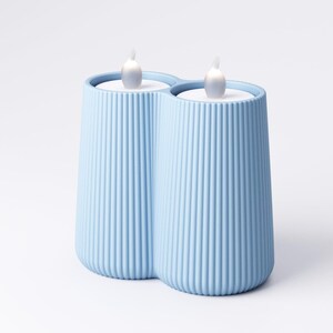 May include: A light blue, ribbed, double candle holder with two white LED flame-shaped lights. The candle holder has a modern design and is made of a solid material. The background is white.
