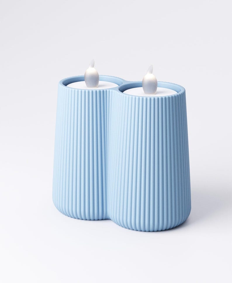 May include: A light blue, ribbed, double candle holder with two white LED flame-shaped lights. The candle holder has a modern design and is made of a solid material. The background is white.