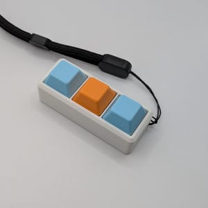 May include: A white rectangular keychain with three square buttons in light blue and orange. A black lanyard is attached to the keychain. The buttons are arranged in a row.