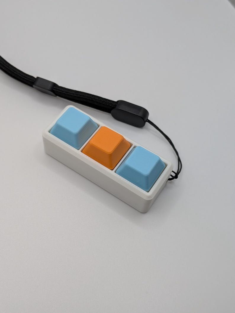 May include: A white rectangular keychain with three square buttons in light blue and orange. A black lanyard is attached to the keychain. The buttons are arranged in a row.