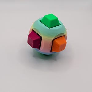 May include: A colorful puzzle ball with a rainbow gradient and six protruding cube-shaped pieces in green, magenta, and orange. The ball is a sphere with a smooth surface and a playful design.