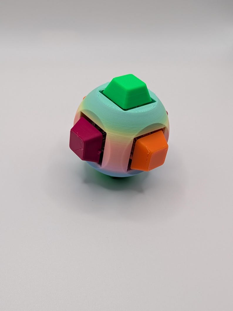 May include: A colorful puzzle ball with a rainbow gradient and six protruding cube-shaped pieces in green, magenta, and orange. The ball is a sphere with a smooth surface and a playful design.