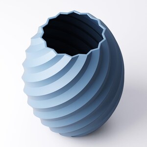 May include: A blue decorative vase with a spiral design. The vase has a textured, ridged surface and a geometric, scalloped rim. The interior is dark blue, contrasting with the lighter blue exterior. The vase is set against a white background.