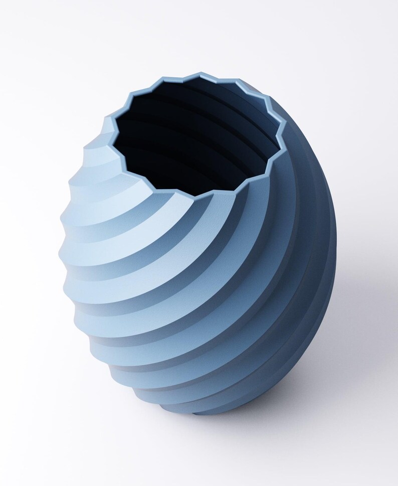 May include: A blue decorative vase with a spiral design. The vase has a textured, ridged surface and a geometric, scalloped rim. The interior is dark blue, contrasting with the lighter blue exterior. The vase is set against a white background.