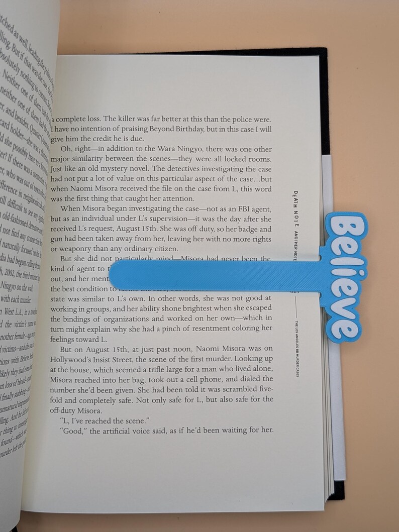 May include: A light blue bookmark with a T-shape design featuring the word Believe in white script lettering. The bookmark is placed inside an open book titled Death Note: Another Note, The Los Angeles BB Murder Cases, with visible text.