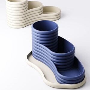 May include: Two modern, sculptural desk organizers. One is off-white, the other is blue. Both have a wavy design with a cylindrical cup and two trays. The organizers are made of a smooth, matte material.