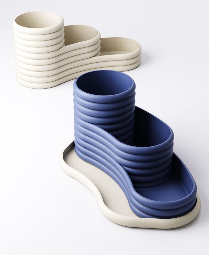 May include: Two modern, sculptural desk organizers. One is off-white, the other is blue. Both have a wavy design with a cylindrical cup and two trays. The organizers are made of a smooth, matte material.