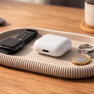 Puede incluir: Una bandeja ovalada beige con borde acanalado contiene un reloj inteligente negro, una funda blanca para auriculares inal&aacute;mbricos, dos anillos y dos monedas. Una taza y un port&aacute;til est&aacute;n en el fondo. La bandeja est&aacute; sobre una superficie de madera.