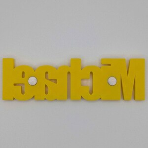 May include: A yellow wooden nameplate with the name "Madison" spelled out in capital letters. The nameplate has two small silver magnets attached to the back. The nameplate is displayed against a white background.