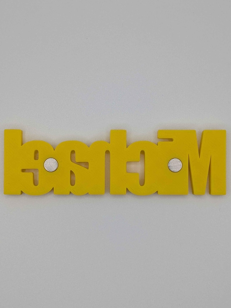 May include: A yellow wooden nameplate with the name "Madison" spelled out in capital letters. The nameplate has two small silver magnets attached to the back. The nameplate is displayed against a white background.