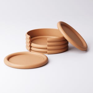 Puede incluir: Un juego de seis posavasos ovalados de color terracota con un borde elevado. Un posavasos est&aacute; separado y otro se ha retirado parcialmente de la pila. Los posavasos est&aacute;n hechos de un material liso y mate.