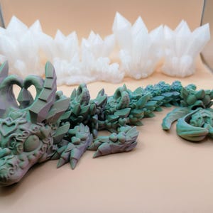 Op de afbeelding: Een gedetailleerd, 3D-geprint drakenbeeldje in groen- en paarstinten, met ingewikkelde details. De draak ligt, met een apart stuk in de buurt. Op de achtergrond staan ​​verschillende witte, kristalachtige structuren.