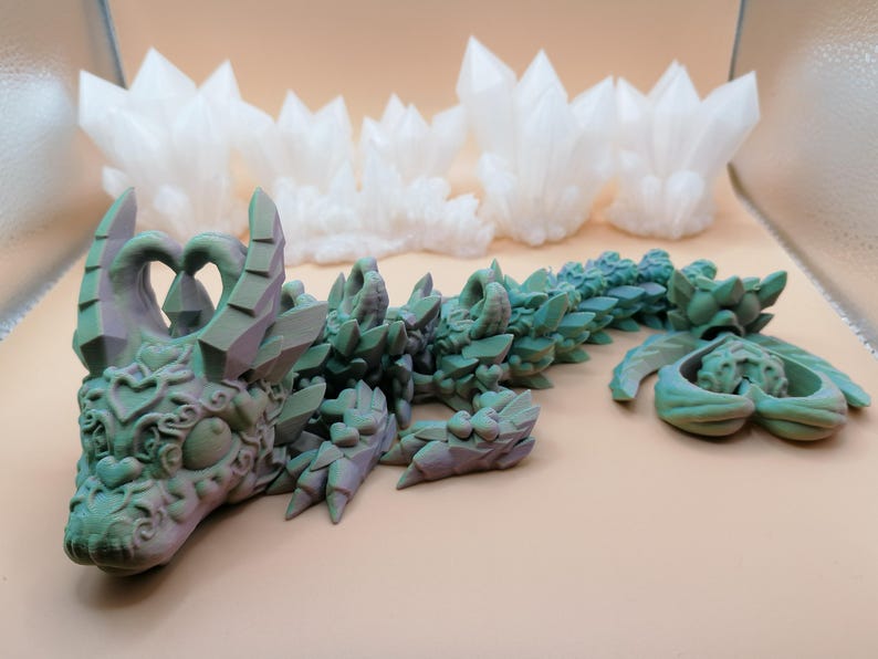 Op de afbeelding: Een gedetailleerd, 3D-geprint drakenbeeldje in groen- en paarstinten, met ingewikkelde details. De draak ligt, met een apart stuk in de buurt. Op de achtergrond staan ​​verschillende witte, kristalachtige structuren.