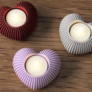 May include: Three heart-shaped candle holders in burgundy, gray, and lavender. Each holder has a ribbed design and holds a white, glowing tea light. The holders are arranged on a wooden surface.