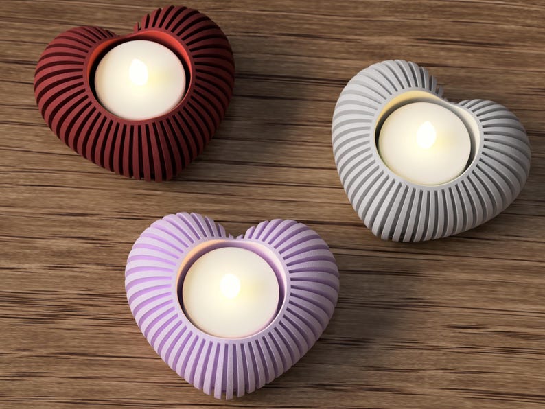 May include: Three heart-shaped candle holders in burgundy, gray, and lavender. Each holder has a ribbed design and holds a white, glowing tea light. The holders are arranged on a wooden surface.
