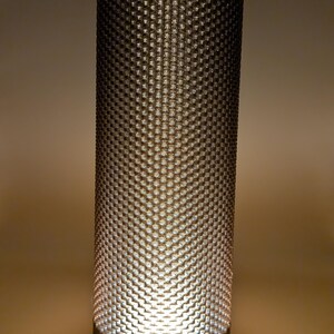 K&ouml;nnte beinhalten: Zylindrische Tischlampe mit strukturiertem, gemustertem Schirm. Der Schirm hat ein sich wiederholendes, gitterartiges Design. Die Lampe strahlt ein warmes, weiches Licht aus und hat einen wei&szlig;en Sockel.