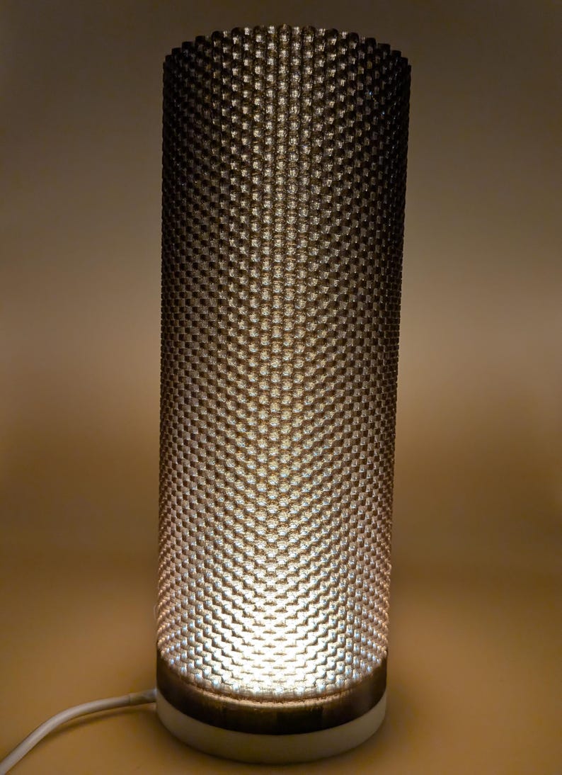 K&ouml;nnte beinhalten: Zylindrische Tischlampe mit strukturiertem, gemustertem Schirm. Der Schirm hat ein sich wiederholendes, gitterartiges Design. Die Lampe strahlt ein warmes, weiches Licht aus und hat einen wei&szlig;en Sockel.