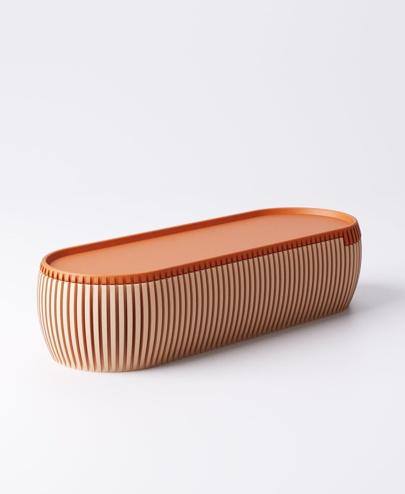 May include: A wooden, oval-shaped storage box with a flat, orange-brown lid. The sides are made of vertical, light-colored wooden slats. The box is approximately 18 inches long.