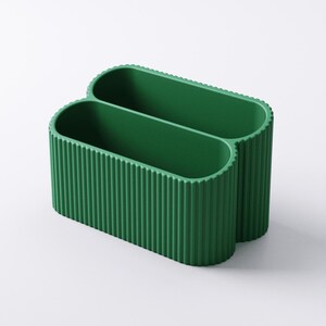 May include: A green, double-compartment desk organizer. The organizer has a ribbed texture on the exterior and a smooth interior. The oval-shaped compartments are connected, creating a unique design. The organizer is made of a solid, opaque material.