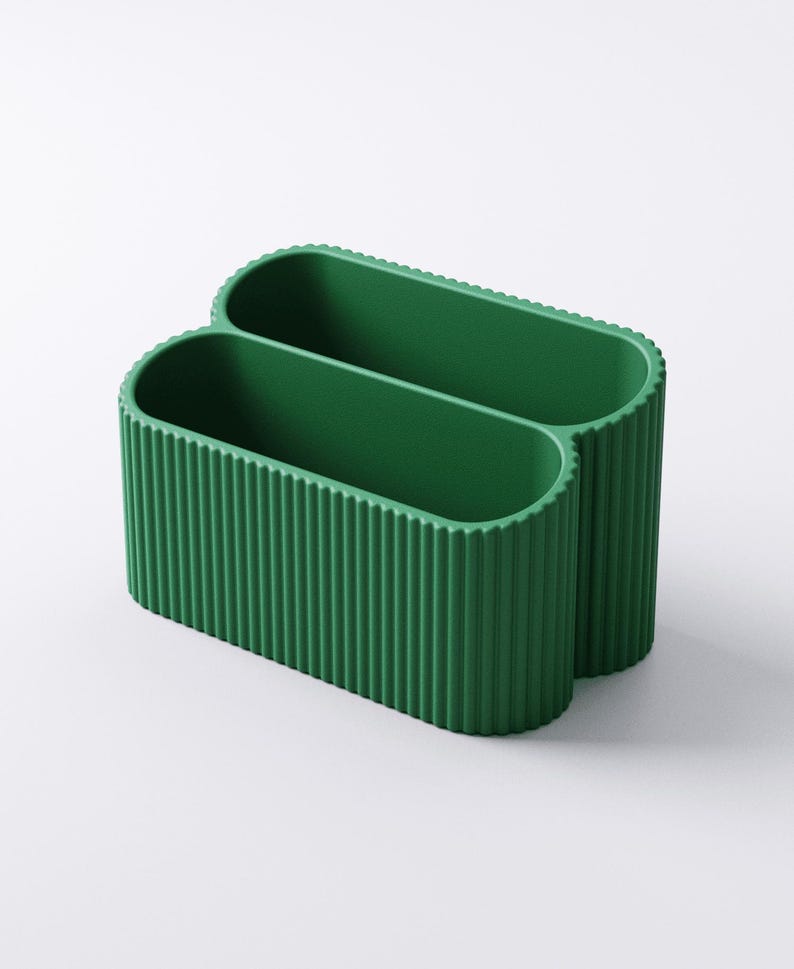 May include: A green, double-compartment desk organizer. The organizer has a ribbed texture on the exterior and a smooth interior. The oval-shaped compartments are connected, creating a unique design. The organizer is made of a solid, opaque material.