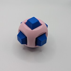 May include: A pink and blue puzzle cube. The cube has a spherical shape with six blue square protrusions. The pink body of the cube has curved indentations between the blue squares. The puzzle is designed to be manipulated and solved.