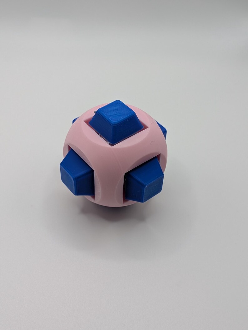 May include: A pink and blue puzzle cube. The cube has a spherical shape with six blue square protrusions. The pink body of the cube has curved indentations between the blue squares. The puzzle is designed to be manipulated and solved.
