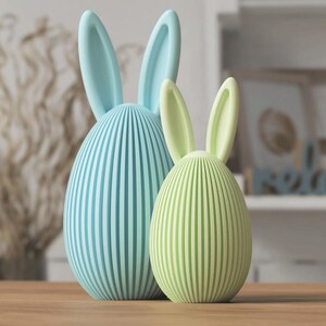 Puede incluir: Dos figuras decorativas de conejos de Pascua. Una es azul claro, la otra verde claro. Ambas tienen patrones de rayas verticales y orejas largas y erguidas. Tienen forma de huevo y est&aacute;n sobre una superficie de madera.