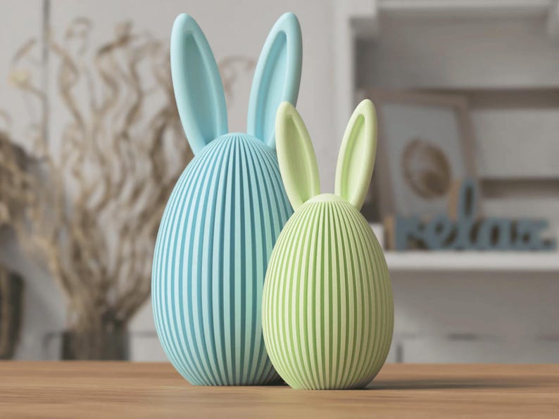 Puede incluir: Dos figuras decorativas de conejos de Pascua. Una es azul claro, la otra verde claro. Ambas tienen patrones de rayas verticales y orejas largas y erguidas. Tienen forma de huevo y est&aacute;n sobre una superficie de madera.