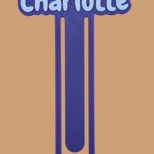 May include: A purple 3D printed bookmark featuring the name Charlotte in light blue lettering. The bookmark has a long, U-shaped design with a rounded bottom and a scalloped border around the name at the top, creating a distinct layered appearance.