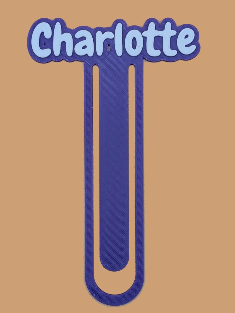 May include: A purple 3D printed bookmark featuring the name Charlotte in light blue lettering. The bookmark has a long, U-shaped design with a rounded bottom and a scalloped border around the name at the top, creating a distinct layered appearance.