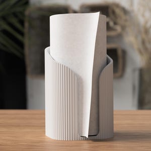 Op de afbeelding: Een moderne, cr&egrave;mekleurige papieren handdoekhouder met een cilindrische basis en verticale geribbelde details. Een rol papieren handdoeken is gedeeltelijk zichtbaar in de houder, met een vel dat uitsteekt. De houder staat op een houten oppervlak.