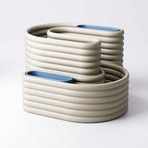 May include: Three beige ceramic containers with a unique, ribbed design. Each has a blue oval insert. The containers are stacked, creating a modern, sculptural display. The measurements are in centimetres.