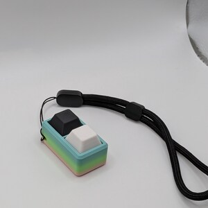 May include: A small, rectangular, two-button device with a black and a white button, set in a pastel-colored frame. The frame transitions from pink to green to blue. A black lanyard is attached to the device.