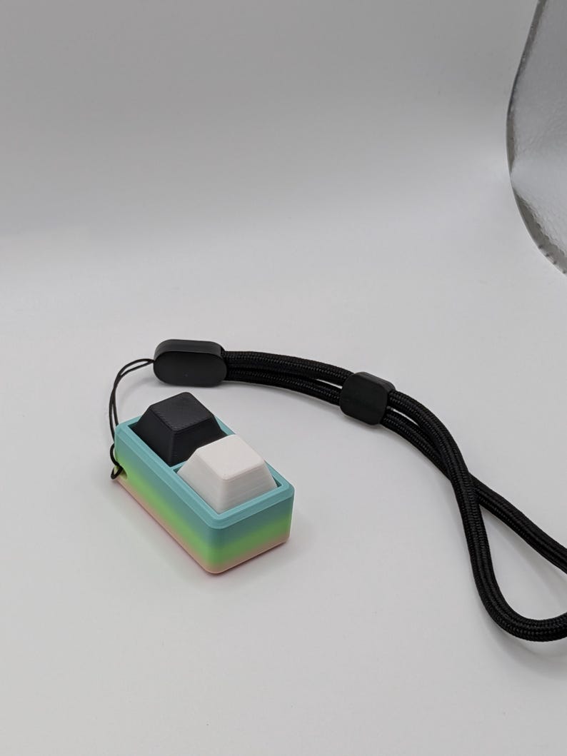 May include: A small, rectangular, two-button device with a black and a white button, set in a pastel-colored frame. The frame transitions from pink to green to blue. A black lanyard is attached to the device.