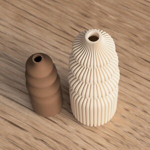 May include: Two decorative vases. One is a light beige color with a spiral design, and the other is a brown, stacked, rounded shape. Both vases have a small opening at the top. The vases are on a wooden surface.