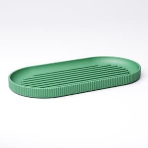 May include: A green, oval-shaped dish drying rack. The rack has a ribbed exterior and several parallel raised lines on the interior. The rack is made of a solid, matte green material and is designed for kitchen use.