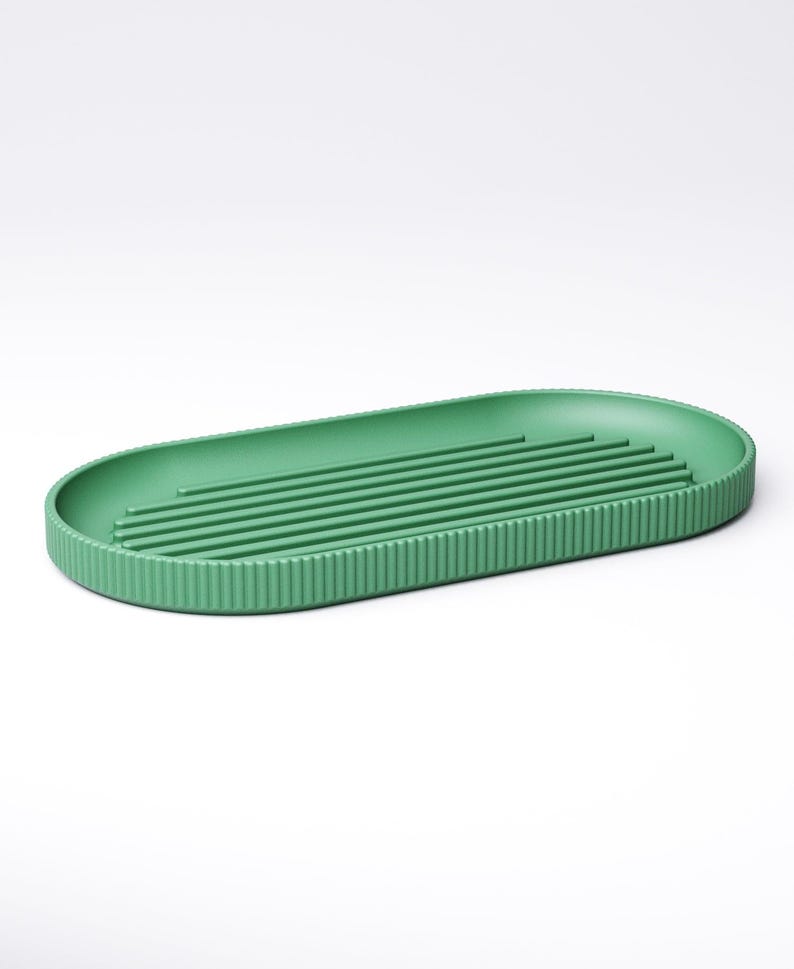 May include: A green, oval-shaped dish drying rack. The rack has a ribbed exterior and several parallel raised lines on the interior. The rack is made of a solid, matte green material and is designed for kitchen use.