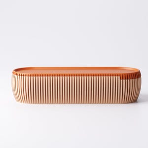 May include: An oval-shaped wooden storage box with a light brown exterior and a darker brown top. The sides are made of vertical wooden slats, creating a textured appearance. The box is designed with a minimalist aesthetic.
