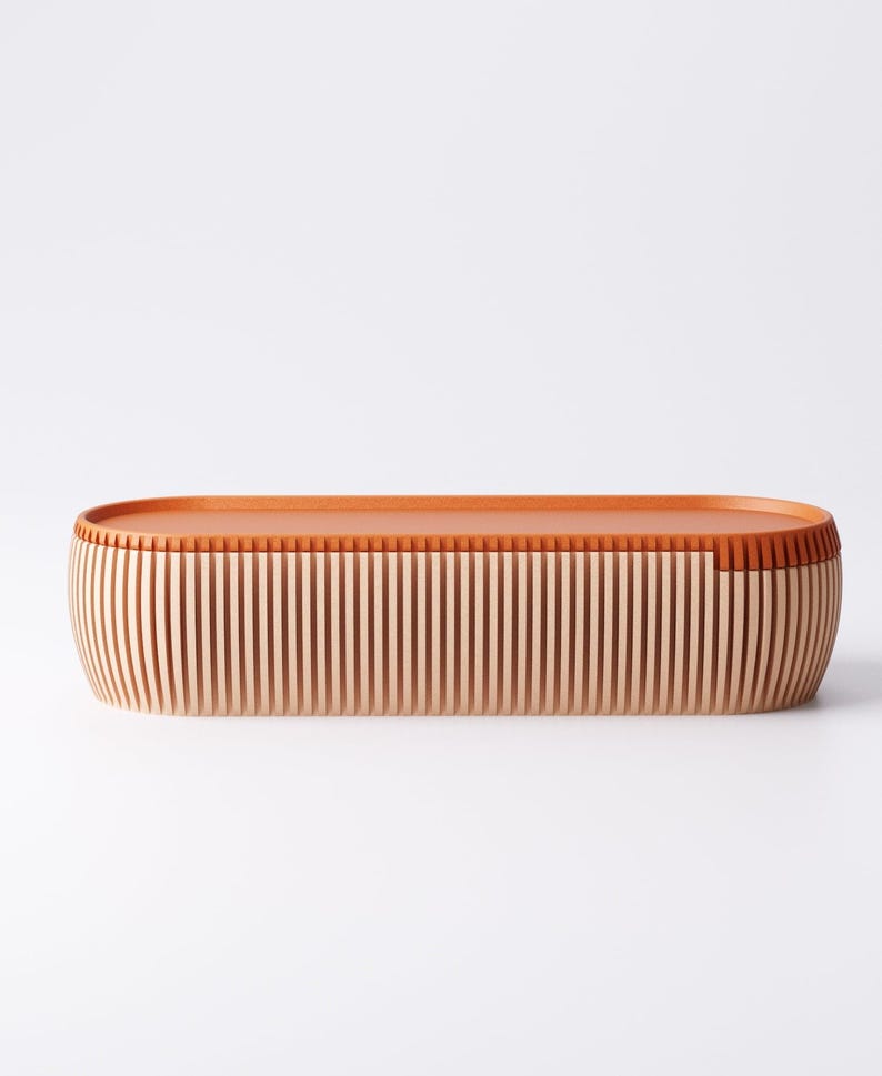 May include: An oval-shaped wooden storage box with a light brown exterior and a darker brown top. The sides are made of vertical wooden slats, creating a textured appearance. The box is designed with a minimalist aesthetic.
