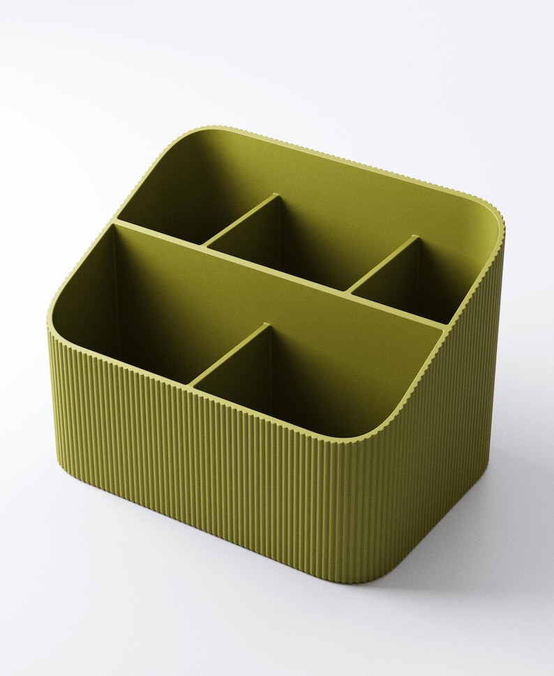 May include: Olive green desk organizer with four compartments. The organizer has a ribbed exterior and rounded corners. The compartments are divided by vertical dividers, creating separate storage spaces for office supplies or other items.