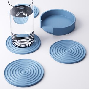 May include: A collection of four pale blue coasters with a concentric circle pattern, a matching holder, and a glass of water. The coasters are made of a solid material and are designed to protect surfaces from water rings.