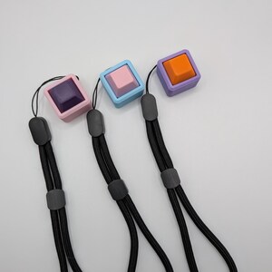 May include: Three square keychains with black lanyards. Each keychain features a different color: purple with a dark purple square, light blue with a pink square, and purple with an orange square. The keychains are on a white background.