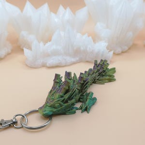 May include: A green and purple dragon-shaped keychain with a silver clasp and ring. The dragon has a textured, detailed design. Several white crystal formations are in the background, set against a light tan surface.
