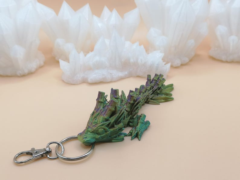 May include: A green and purple dragon-shaped keychain with a silver clasp and ring. The dragon has a textured, detailed design. Several white crystal formations are in the background, set against a light tan surface.