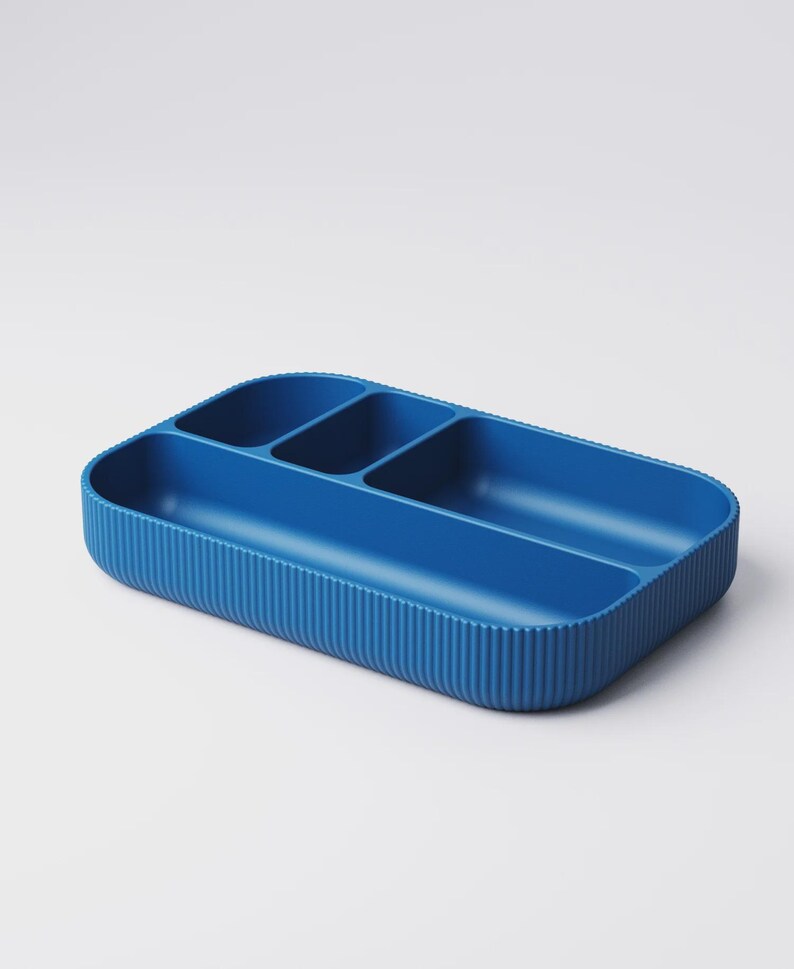 May include: A blue rectangular organizer with four compartments. The exterior features vertical ribbed detailing, while the interior has smooth, curved dividers. This storage container is designed for organizing small items.
