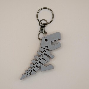 May include: A gray dinosaur skeleton keychain with a metal clasp and ring. The dinosaur is a stylized representation of a T-Rex, with articulated segments. The keychain is made of a 3D-printed material.