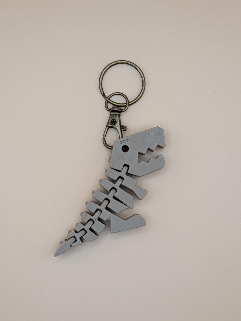 May include: A gray dinosaur skeleton keychain with a metal clasp and ring. The dinosaur is a stylized representation of a T-Rex, with articulated segments. The keychain is made of a 3D-printed material.